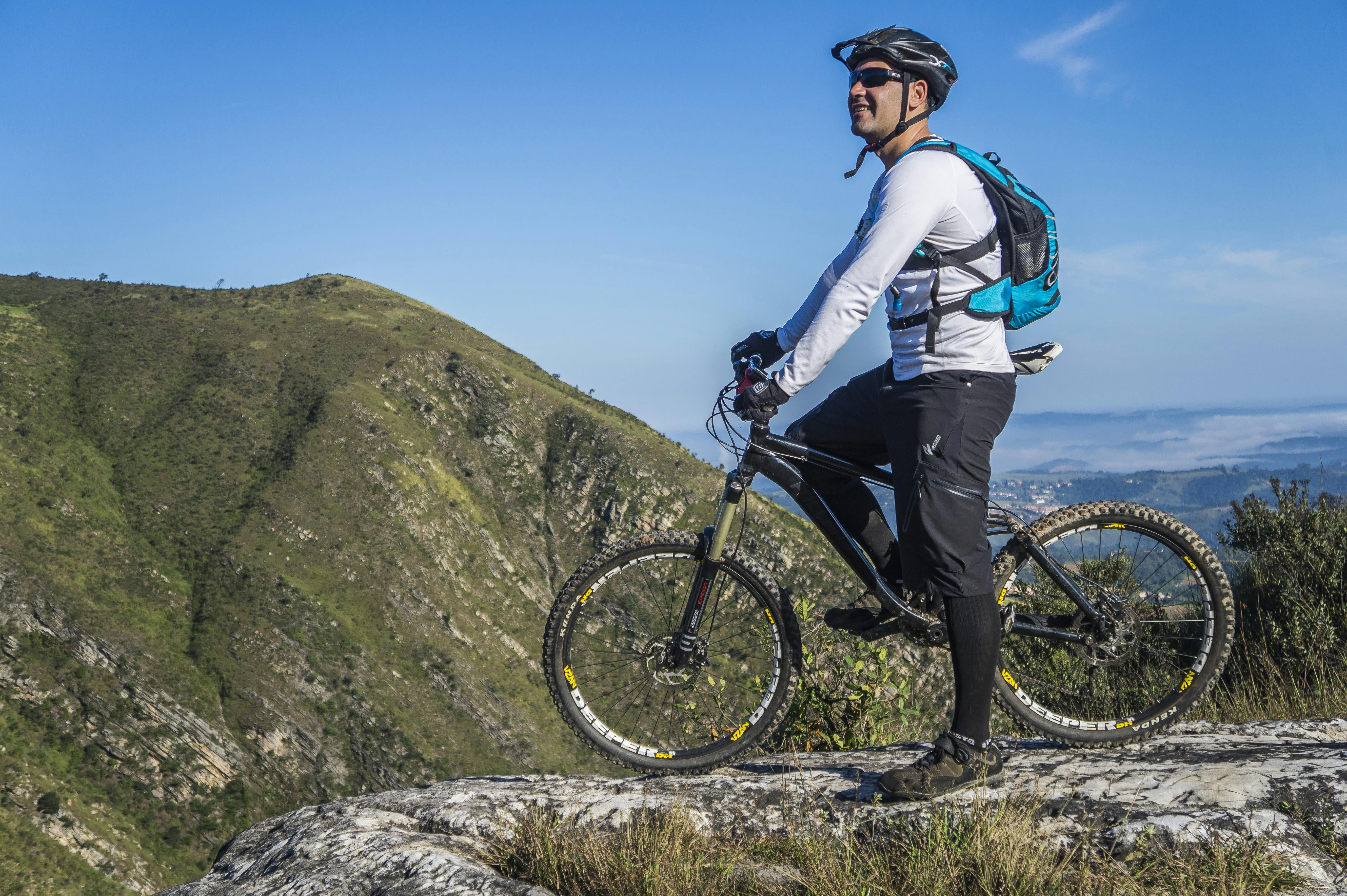 A person in biking gear stands with a mountain bike on a rocky ledge, overlooking rolling green hills under a blue sky.